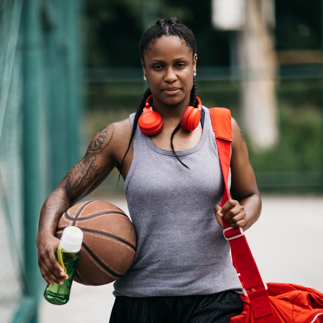 Woman holding a basketball and a water bottle, wearing a gray tank top and red headphones, with a red bag over her shoulder.