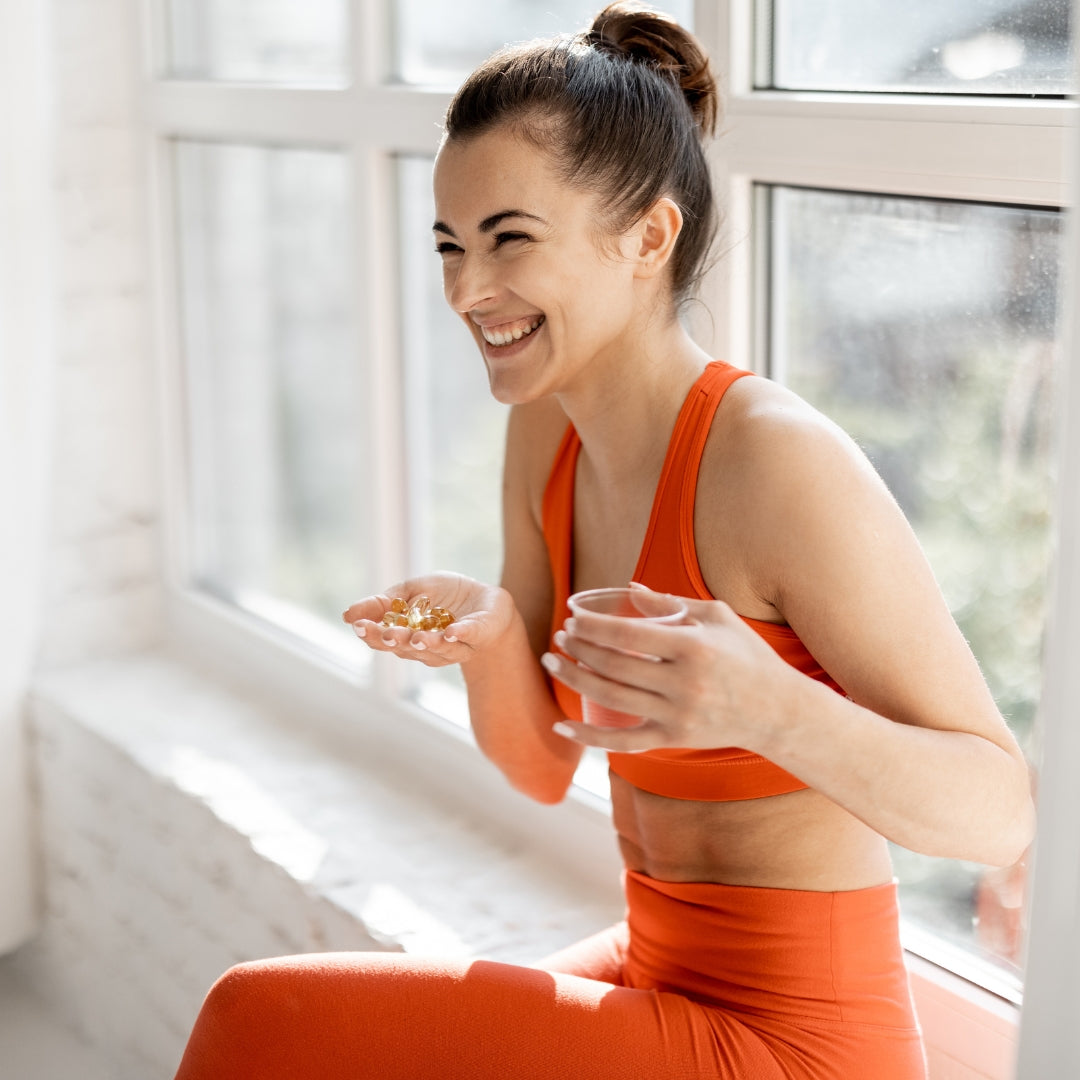 Woman in orange athletic wear holding a supplement and a glass of water by a window.
